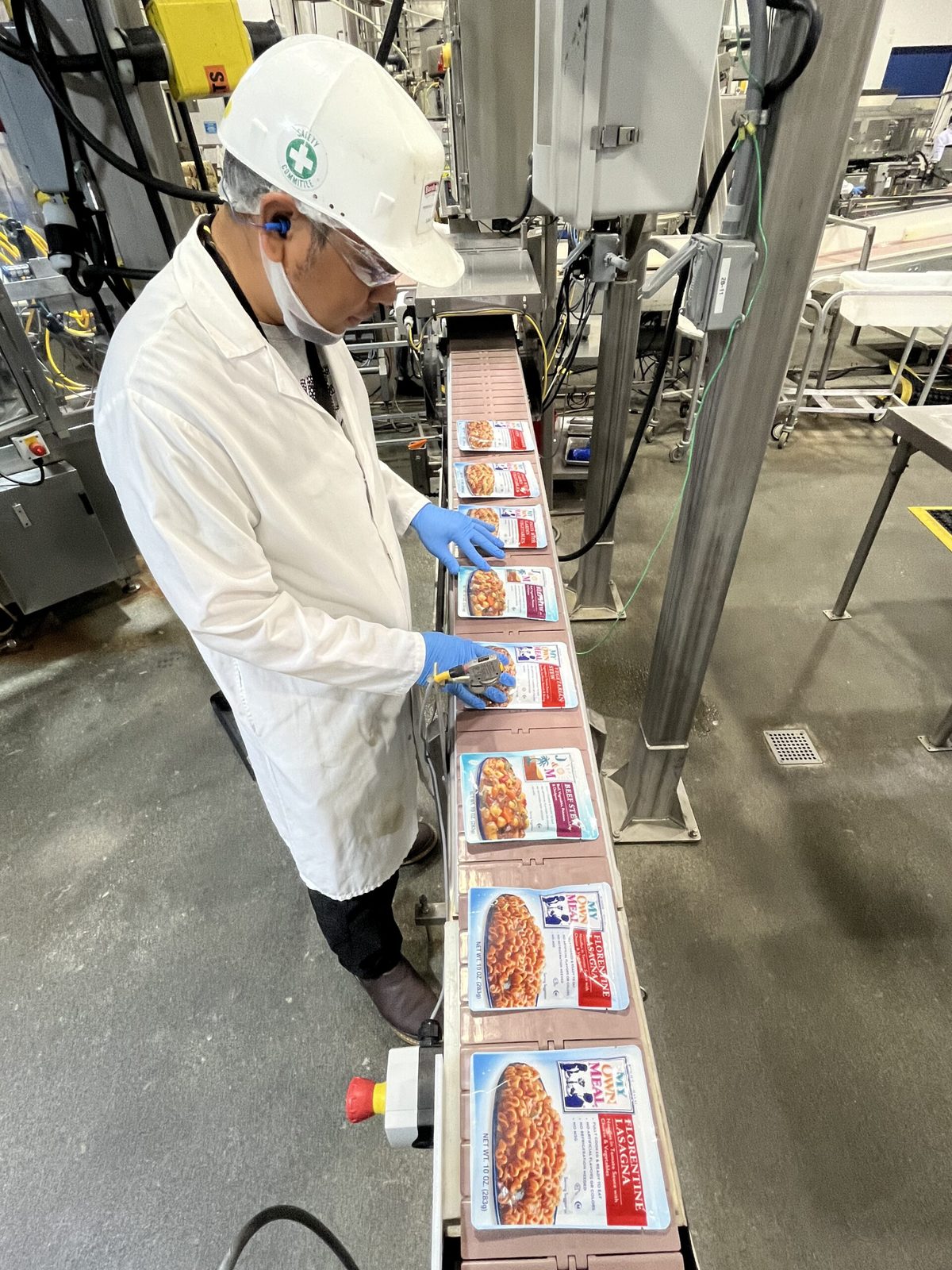 A worker in a hard hat and gloves inspecting packaged meal pouches on a production line