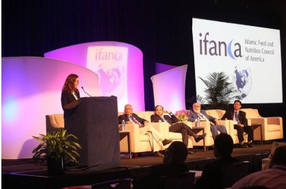 Mary Anne Jackson speaking at a podium beside a panel seated on stage at IFANCA's 19th Annual International Halal Food Conference