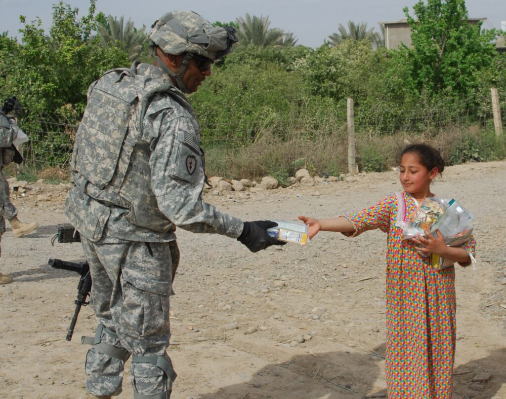 A soldier in uniform hands a packaged meal to a young girl on a dirt road